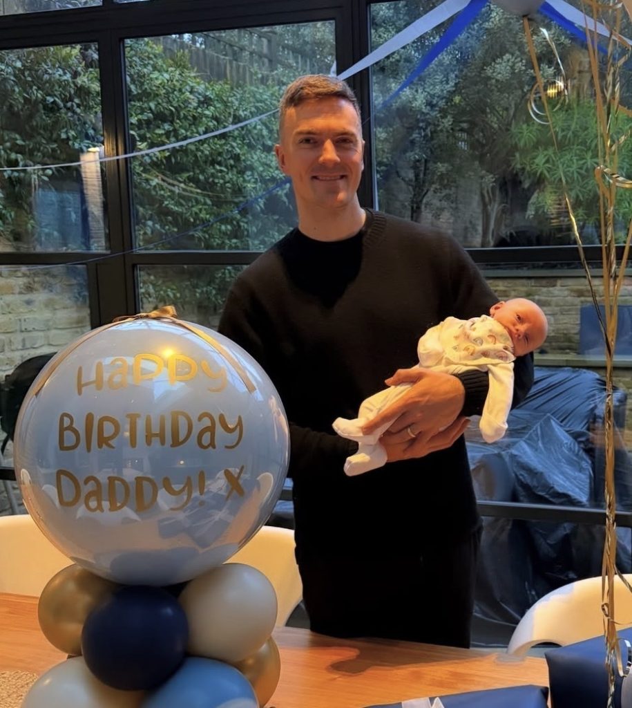 Peloton instructor Ben Alldis holding his sone next to balloons that say Happy Birthday Daddy