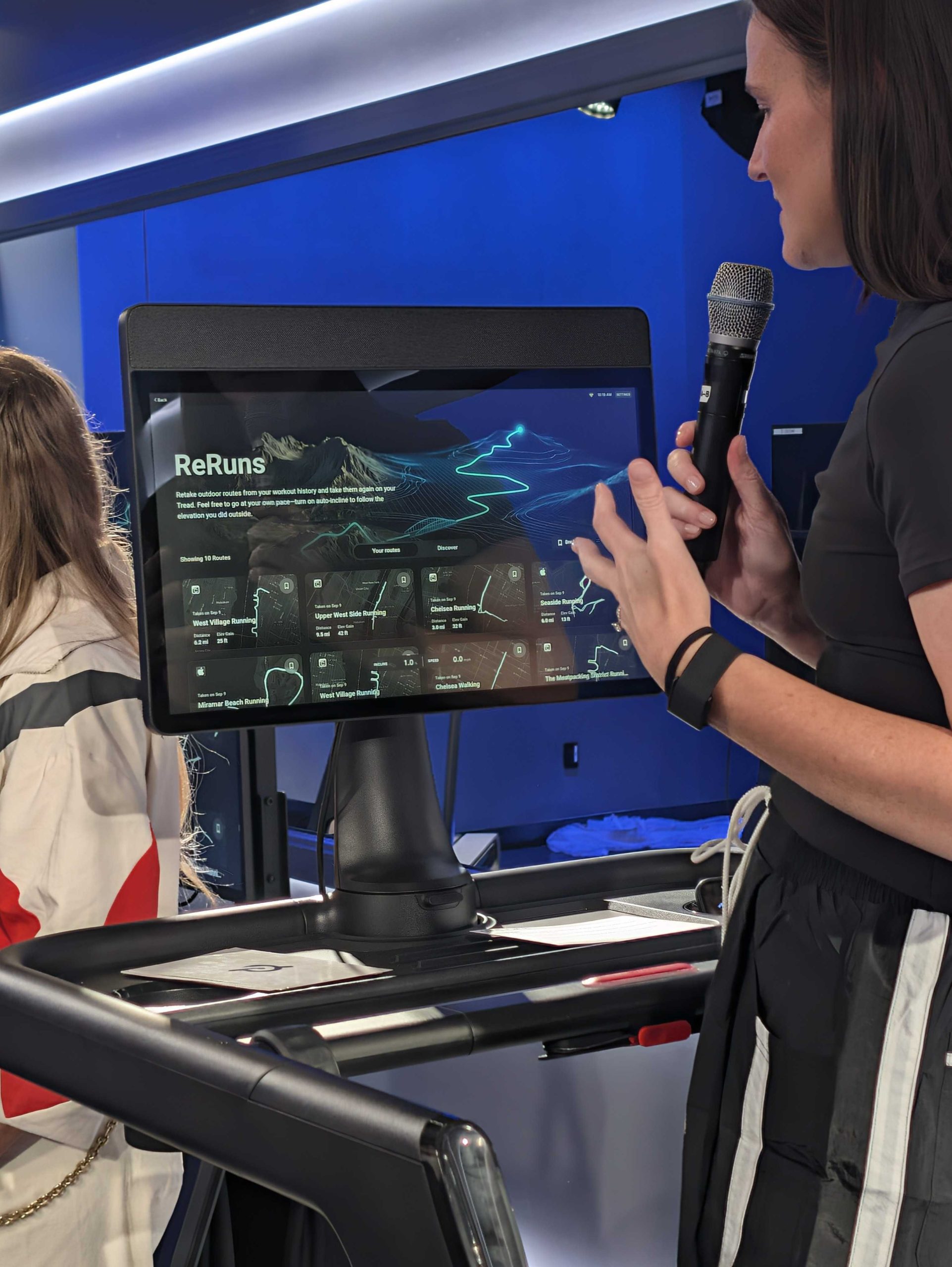 image showing new ReRuns feature on the new Peloton Tread Vision. Meg is standing on the Tread with a microphone explaining the feature.
