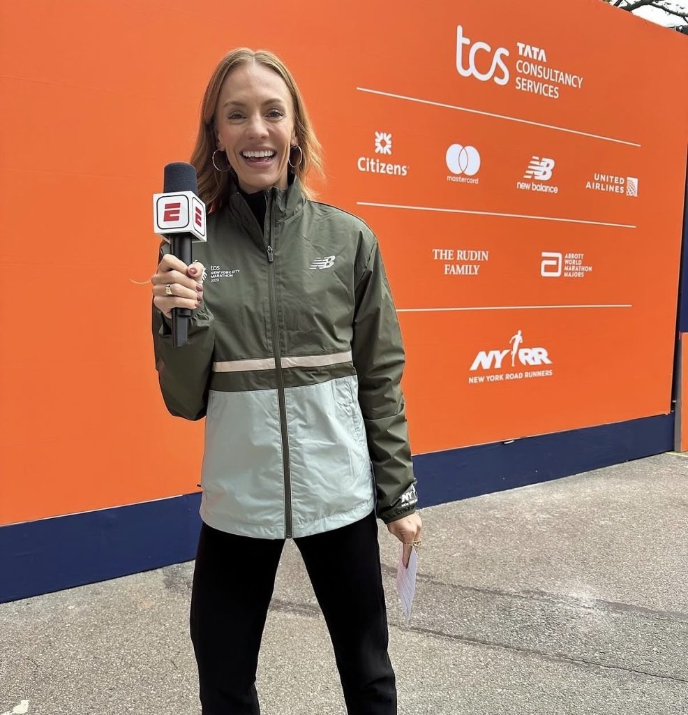 Becs Gentry NYC Marathon reporter Becs Gentry holding an ESPN microphone in front of the TCS New York City Marathon sponsor wall in 2023